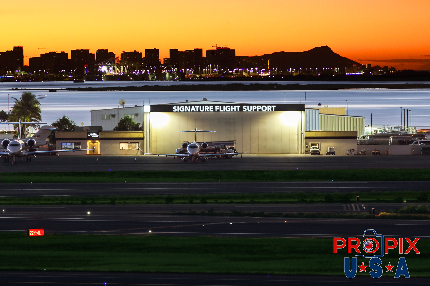 Signature FBO (Private aviation support) at The Honolulu Airport on Oahu Hawaii. Diamondhead and the Waikiki beach area are seen in the background. The Iconic Hilton Hawaiian village tower is seen as a colorful illuminated sliver overlooking the coast.