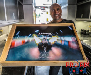 NHRA Top Fuel driver and World Champion Antron Brown holding autographed / signed print of his dragster car launching at night.