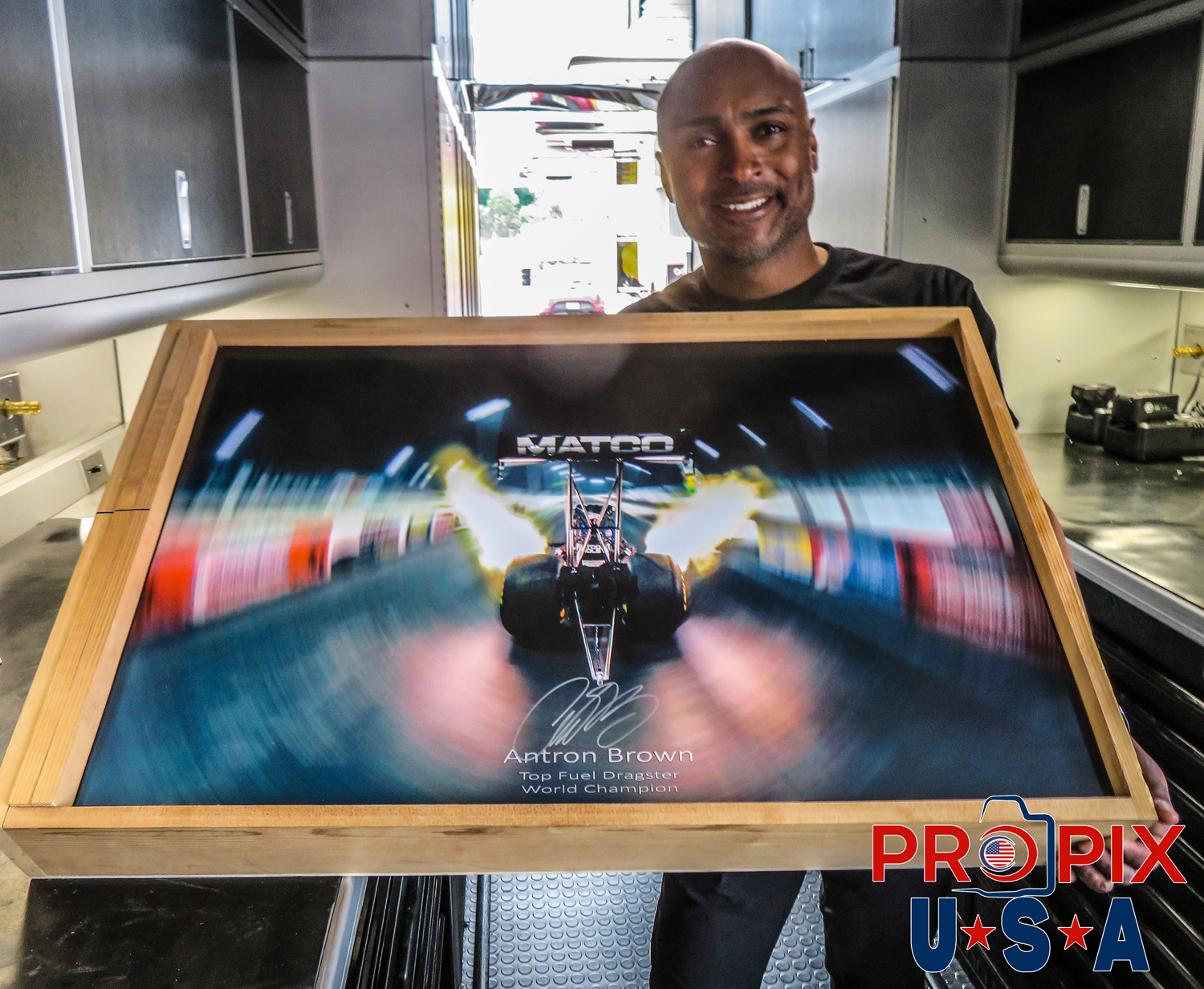 NHRA Top Fuel driver and World Champion Antron Brown holding autographed / signed print of his dragster car launching at night.