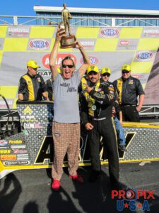 Nicko McBrain from the band Iron Maiden snags then hoists the NHRA Top Fuel Winners trophy from Tony Schumacher.
